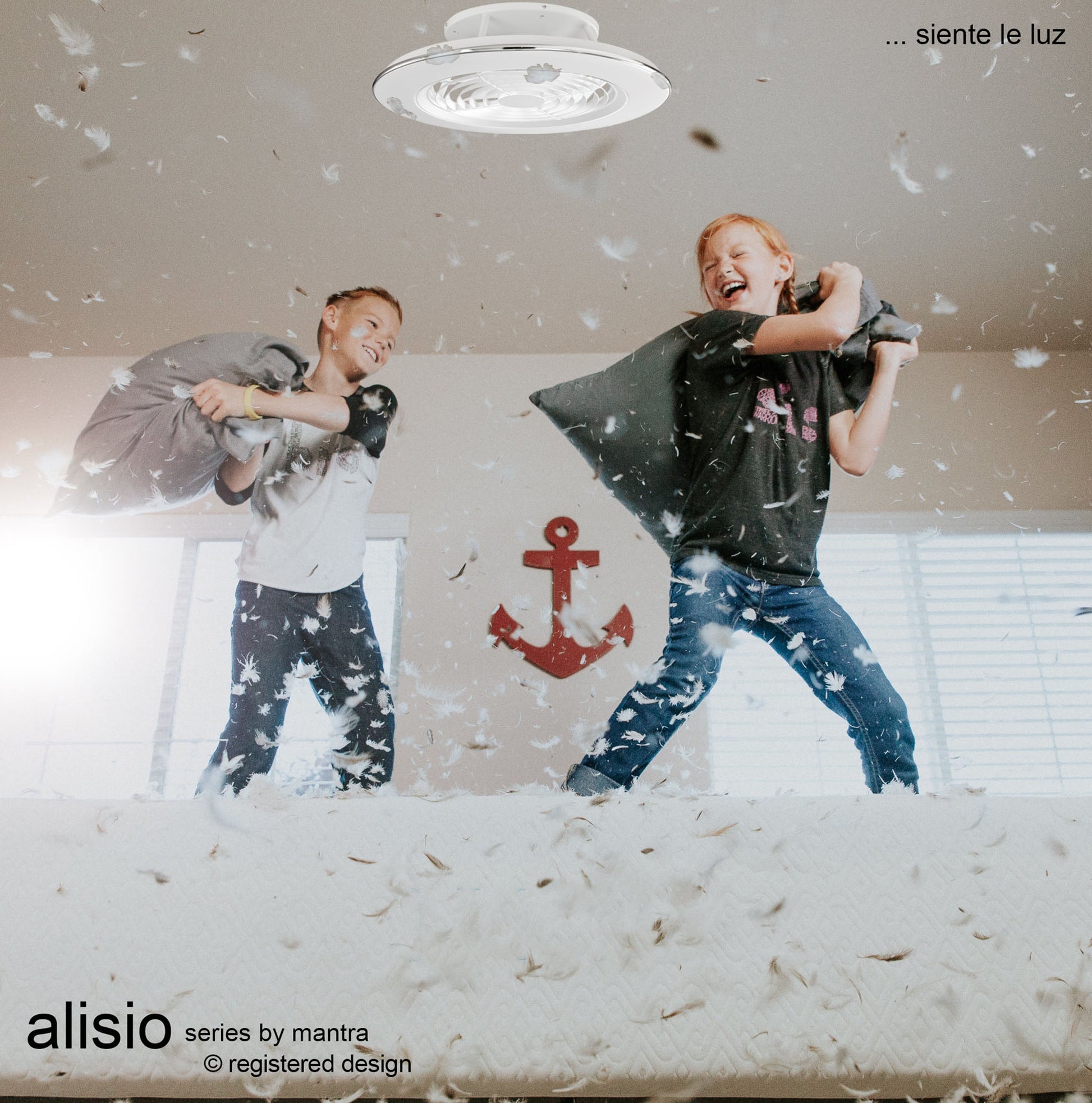 Two children are joyfully engaged in a pillow fight on a bed, causing feathers to fly all around. Above them, an Alisio White Ceiling Fan with Light from Ceiling Fans UK illuminates the room with a cheerful glow. A red anchor decoration is on the wall behind them. The text reads "Alisio series by Mantra … siente la luz.