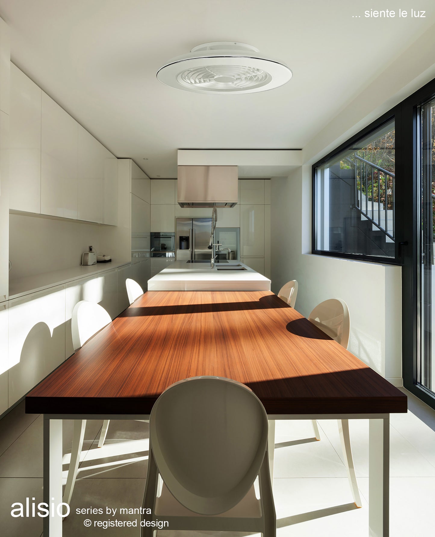 A modern kitchen with white cabinetry and a long wooden dining table surrounded by white chairs. Large windows on the right wall allow natural light to flood the room. Centrally located above the table is a sleek Alisio White Ceiling Fan with Light from Ceiling Fans UK, featuring a 63cm, 70W LED dimmable ceiling light and a built-in 35W DC reversible fan, all in a white finish and controlled via remote or app. Text reads "alisio series by mantra" and "... siente le luz.