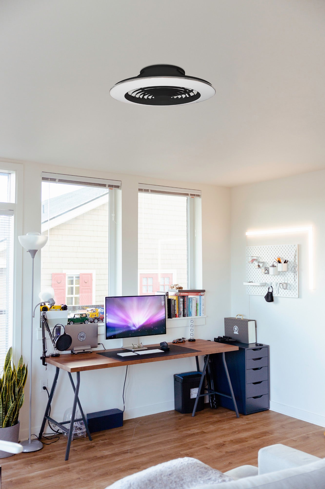 A modern home office with a wooden desk holding a computer monitor, keyboard, and speakers. There are shelves and a pegboard on the wall above the desk, and large windows providing natural light. The sleek Ceiling Fans UK Alisio XL Black Ceiling Fan with Light 73.5cm 95W LED Dimmable Ceiling Light & 58W DC Reversible Fan in Black Finish features an LED dimmable ceiling light for optimal illumination and comes complete with remote control, APP & Alexa/Google Voice Control (5900lm).