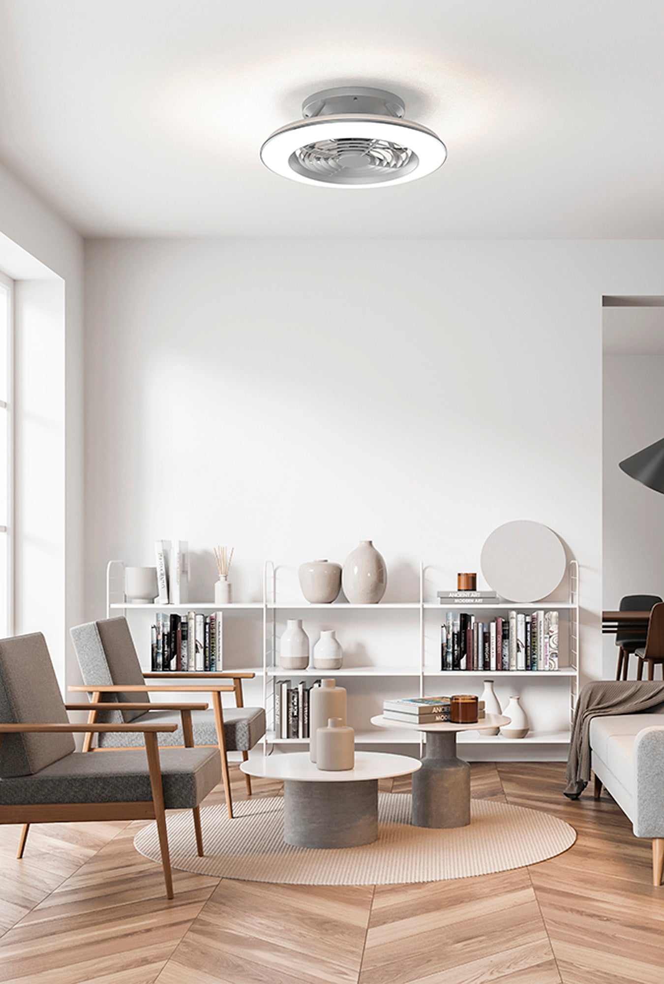 A modern, bright living room with natural light streaming through a large window. The room boasts two gray armchairs, a small white circular coffee table, and minimalist decor. Centered above is the Alisio Mini Silver Ceiling Fan with Light by Ceiling Fans UK, featuring a built-in 30W DC reversible fan and 70W LED dimmable lighting that brightly illuminates the space. Bookshelves are adorned with books and decorative items, enhancing the room's appeal.

