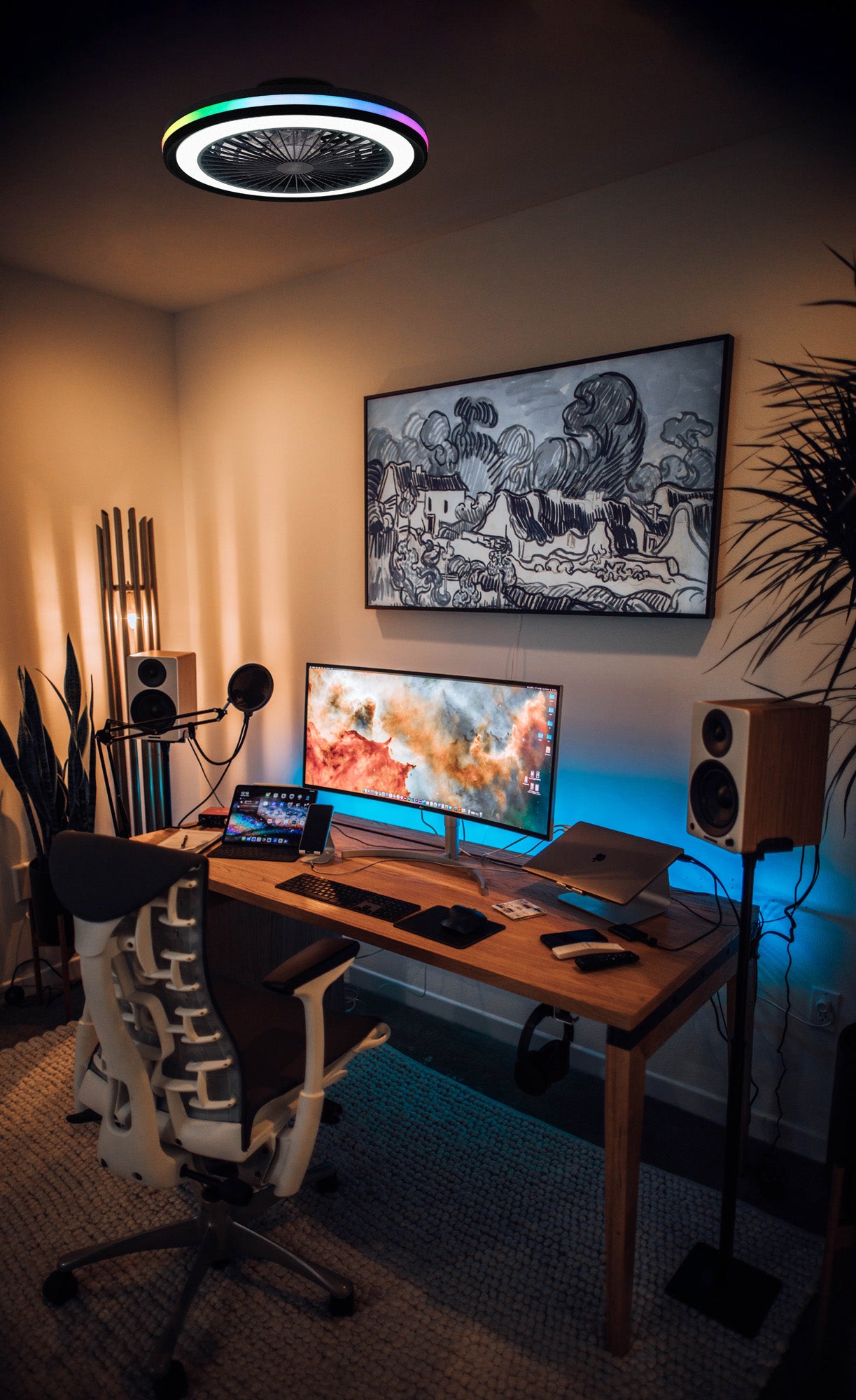 A modern home office setup featuring a wooden desk with a widescreen monitor, a laptop, and various peripherals. A stylish chair is in front of the desk, illuminated by an LED dimmable light. The room is decorated with large framed artwork, ambient lighting, several plants, and even boasts the Ceiling Fans UK Gamer Small Black Ceiling Fan with Light 47cm 40W LED Dimmable White/RGB Ceiling Light With Built-In 20W DC Reversible Fan, controlled via remote control.